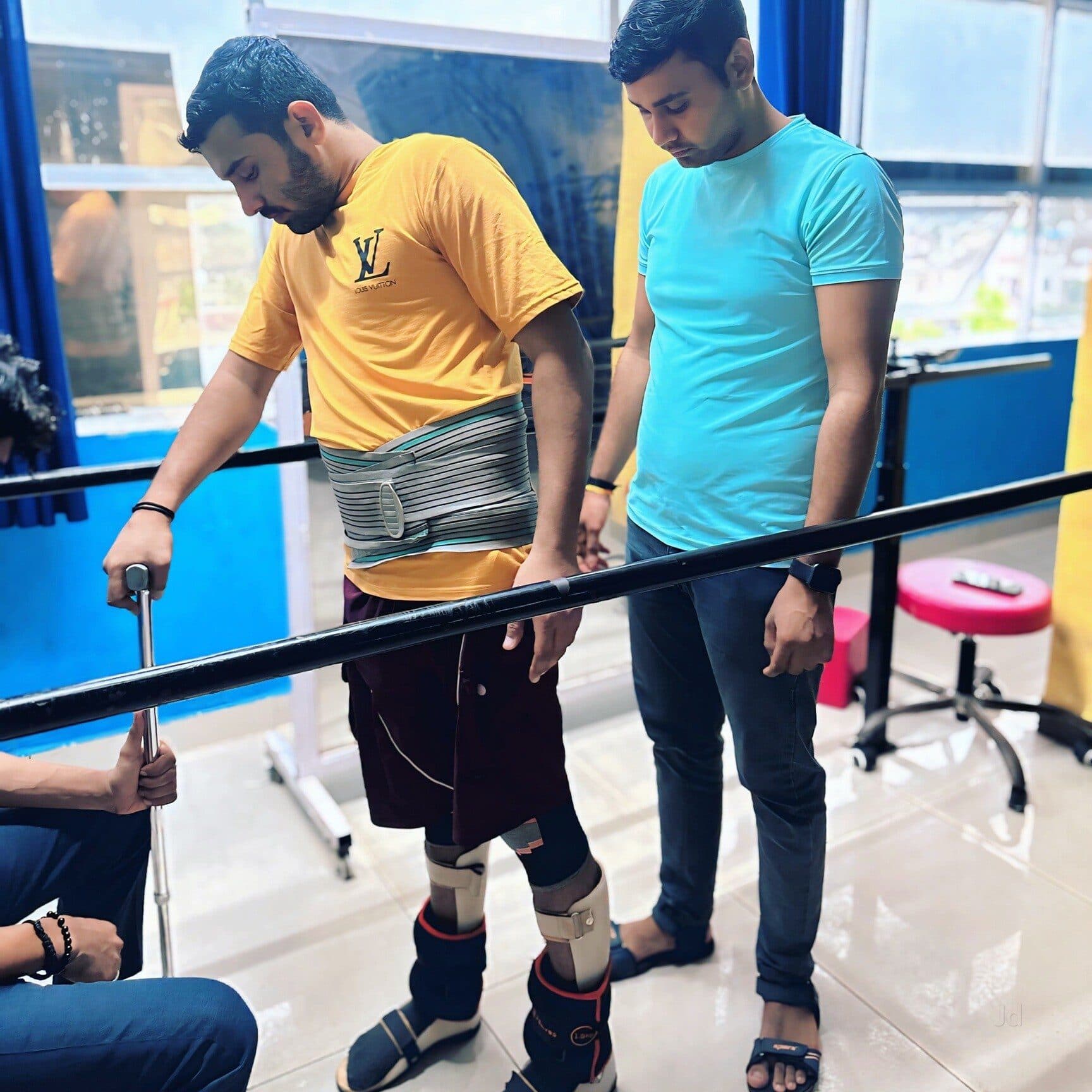 A patient wearing a lumbar belt and leg braces walking between the parallel bars at Quick Walk, a physiotherapist guarding from behind